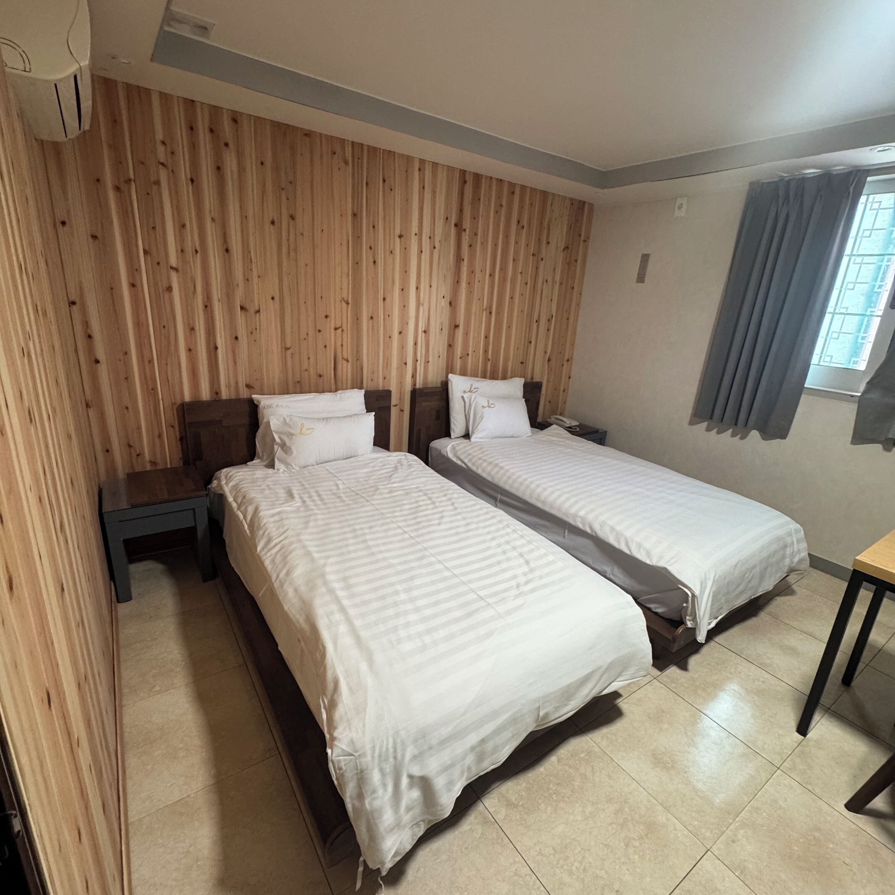 A hotel room with two single beds, a wooden headboard, a small table, and a window with curtains.