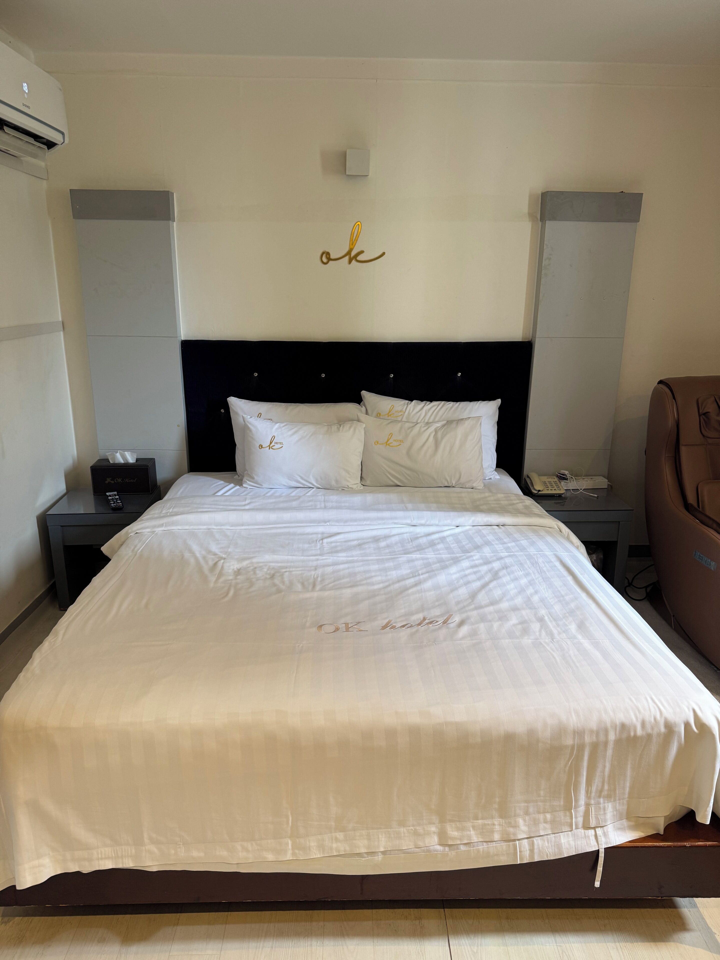 Une chambre d’hôtel avec un grand lit, une tête de lit, une table de chevet avec une télécommande, une lampe de chevet et un fauteuil en cuir marron.