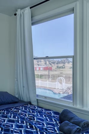 A bedroom with a bed, a window with a view of a fenced area and a red building.