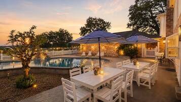 Uma área de terraço ao ar livre com piscina, mesas e cadeiras brancas, e guarda-sóis.