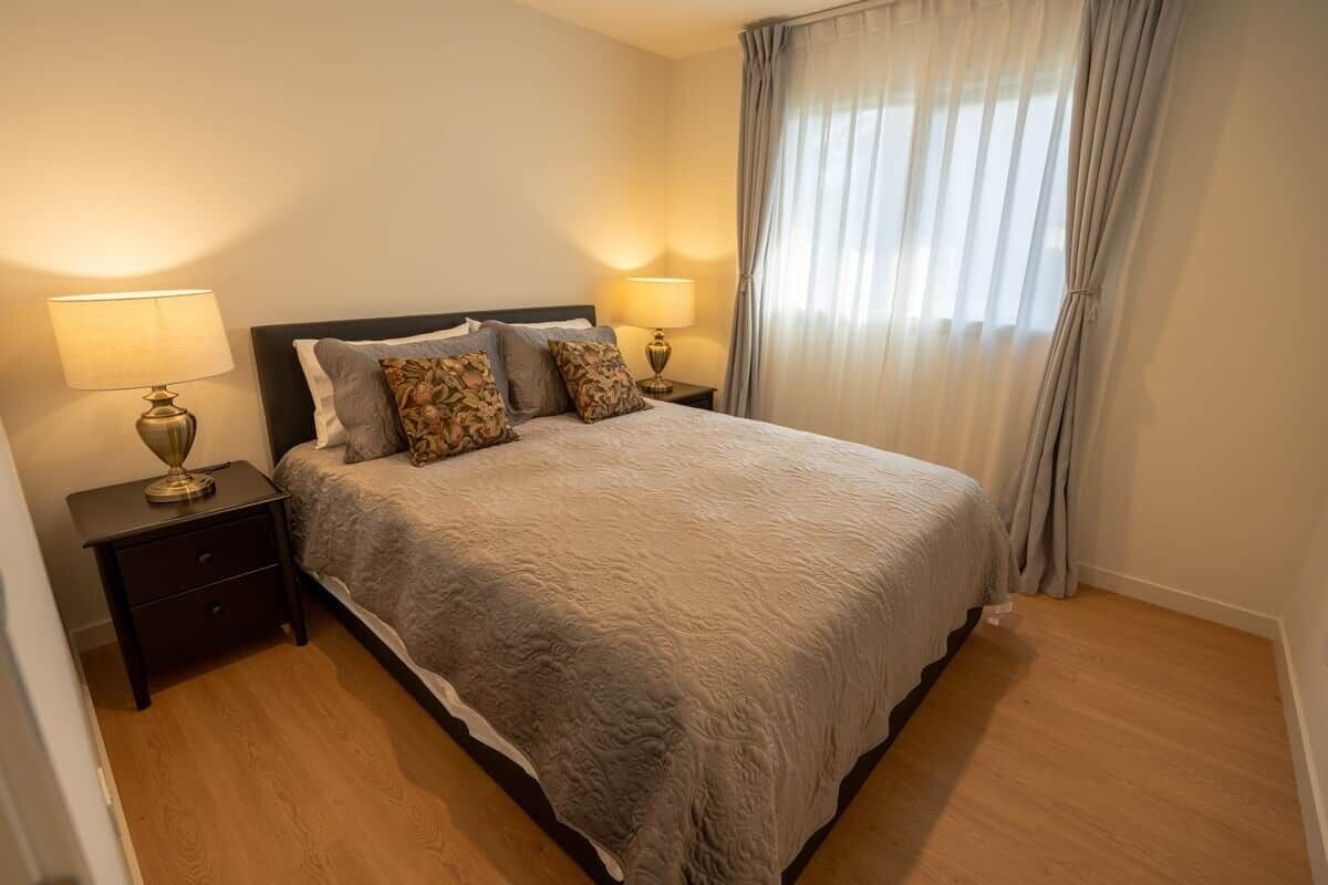 A bedroom with a bed, two bedside tables with lamps, a window with curtains, and a wooden floor.