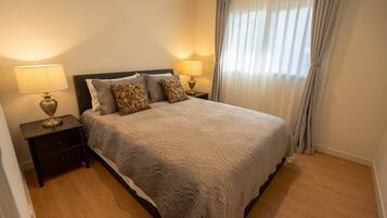 A bedroom with a bed, two bedside tables with lamps, a window with curtains, and a wooden floor.