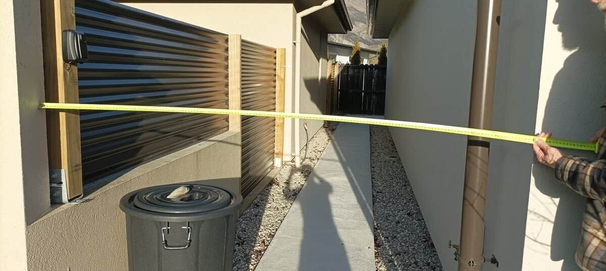 A narrow outdoor corridor with a yellow measuring tape, a trash can, and a window with blinds.