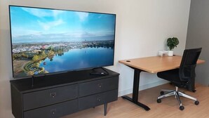 A modern room with a flat-screen TV, a wooden desk, a black office chair, a small potted plant, and a stainless steel refrigerator.