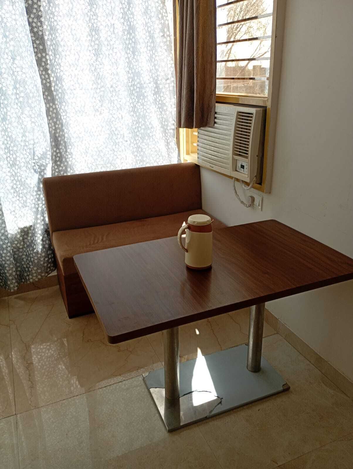 Una habitación con un banco de madera, una mesa con una taza, una ventana con cortinas y un equipo de aire acondicionado.