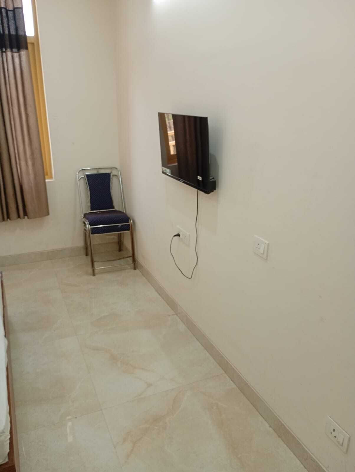 Una habitación con piso de baldosas, una silla, un televisor de pantalla plana montado en la pared y una ventana con cortinas.