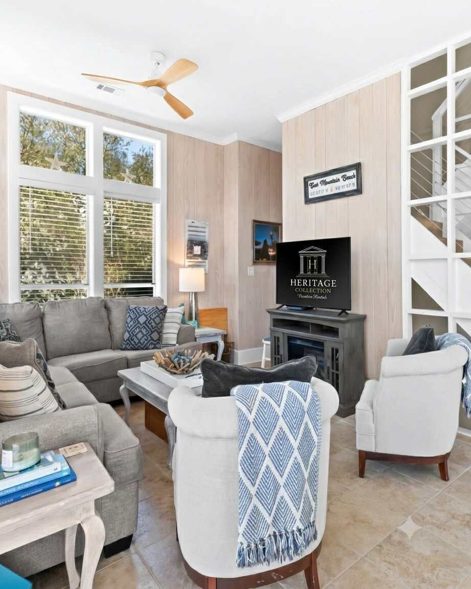 A modern living room with a sofa, armchairs, a coffee table, and a television. There is a staircase to the right and a window with blinds.