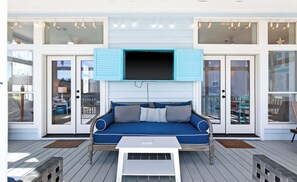 A covered outdoor seating area with a blue sofa, a television mounted above, and a wooden deck. - My Blue Sky - 4 Bikes, Fire Pit & Porch Paradise (Santa Rosa Beach)
