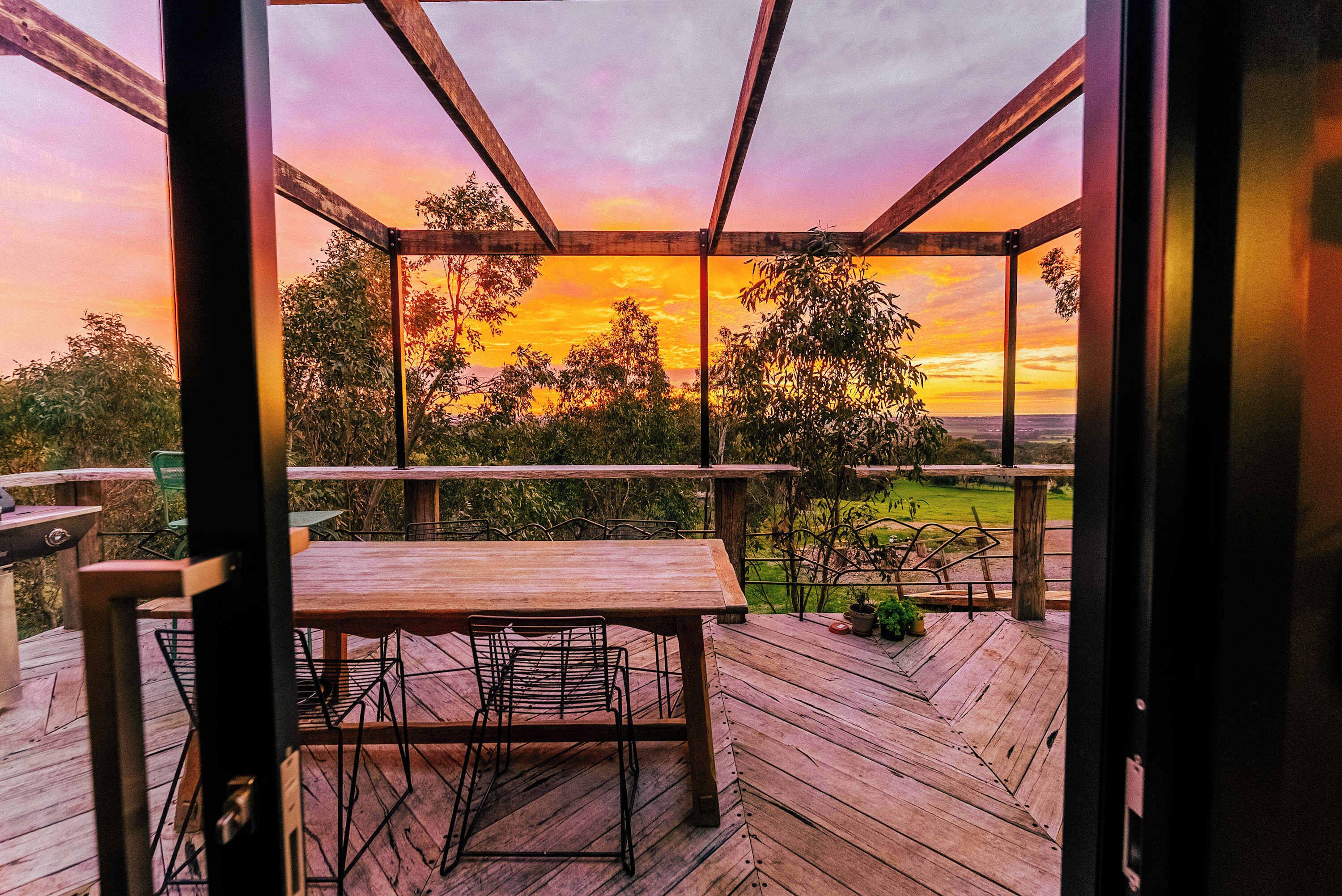 An outdoor wooden deck with a table and chairs, overlooking a landscape with trees and a sunset.