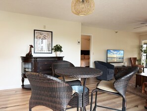A living room with a dining area, featuring wicker furniture, a large painting, and a view into another room. - Sandpiper Key 2BR Bay Views & Heated Pools (Englewood)