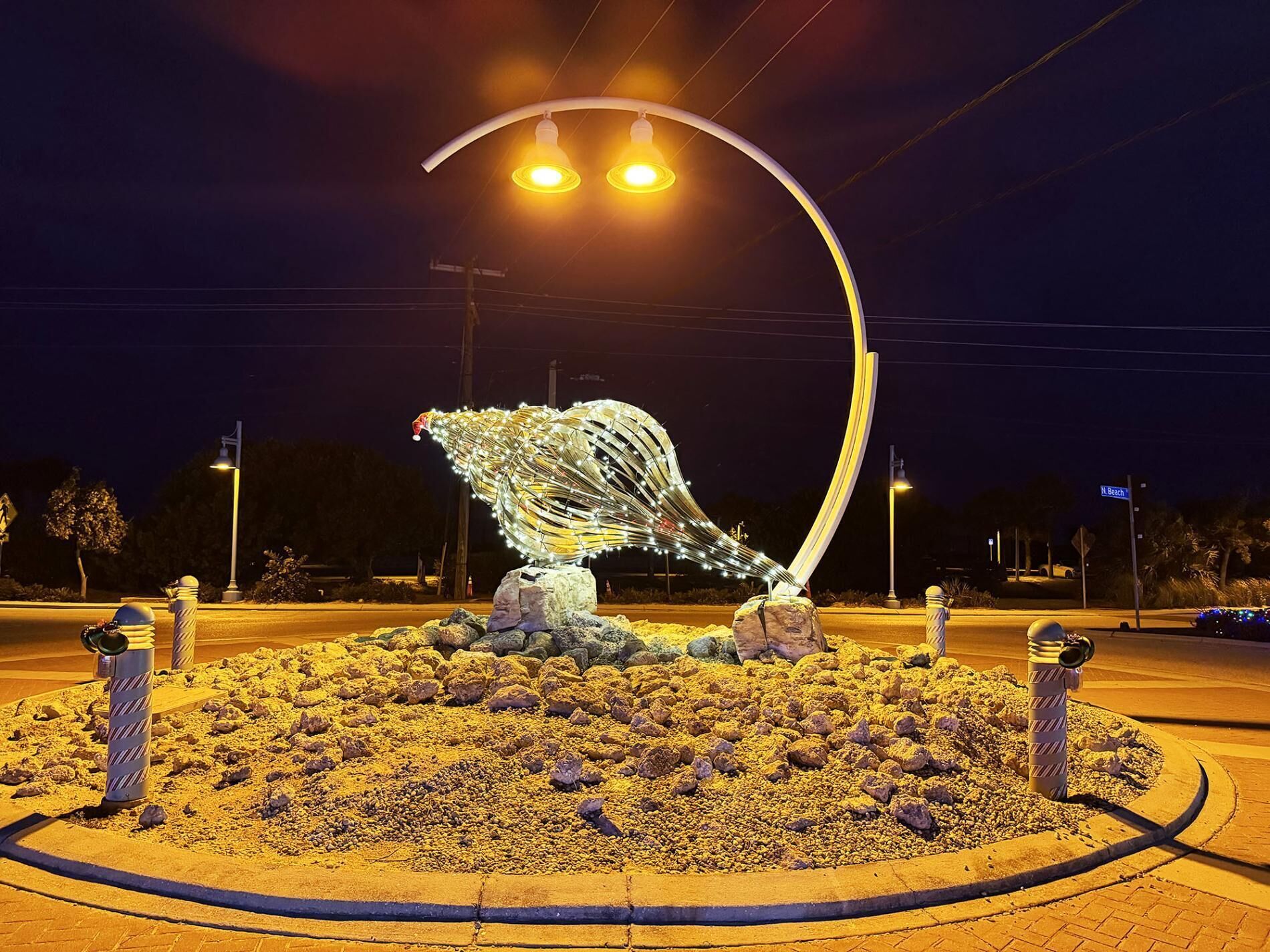 A nighttime street scene with a decorative sculpture in the center, surrounded by a circular gravel area and street lamps.