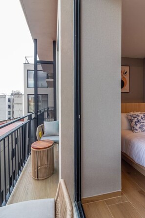 Ein Balkon mit Sitzgelegenheit, ein Bett mit gemustertem Kopfkissen und Blick auf die Stadt.
