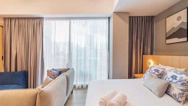 Hotellihuone, jossa on sänky, sohva, ikkuna verhoilla ja seinälle ripustettu taideteos.