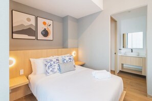 Une chambre d’hôtel comprenant un lit, des tables de chevet, une commode, une salle de bain avec un lavabo et un miroir, et deux œuvres d’art encadrées accrochées au mur.