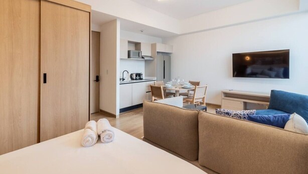 Ein Hotelzimmer mit Bett, Sofa, Esstisch mit Stühlen, Küchenzeile und einem an der Wand befestigten Flachbildfernseher.