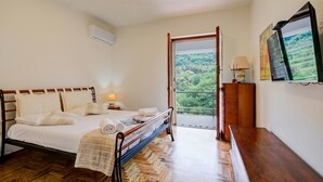 Une chambre à coucher comprenant un lit, une table de chevet, une lampe, une commode et donnant sur un balcon agrémenté de verdure.
