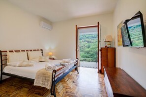 Ein Schlafzimmer mit einem Bett, einem Nachttisch, einer Lampe, einer Kommode und Blick auf einen Balkon mit Grünpflanzen.