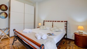 Un lit bien fait, avec du linge de lit blanc et des oreillers, une tête de lit en bois, une table de chevet avec une lampe et une armoire avec un miroir.
