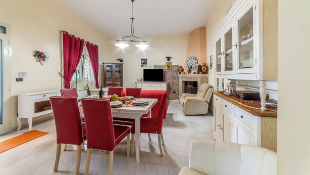 Uma sala de jantar com uma mesa posta para quatro pessoas, cadeiras vermelhas, um armário branco com portas de vidro e uma televisão ao fundo.