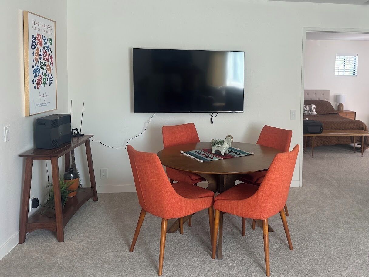 A dining area with a round table, six orange chairs, a flat-screen TV mounted on the wall, and a small wooden side table with a plant.