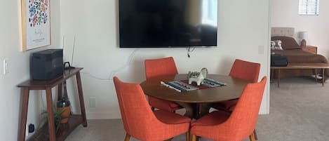 A dining area with a round table, six orange chairs, a flat-screen TV mounted on the wall, and a small wooden side table with a plant.