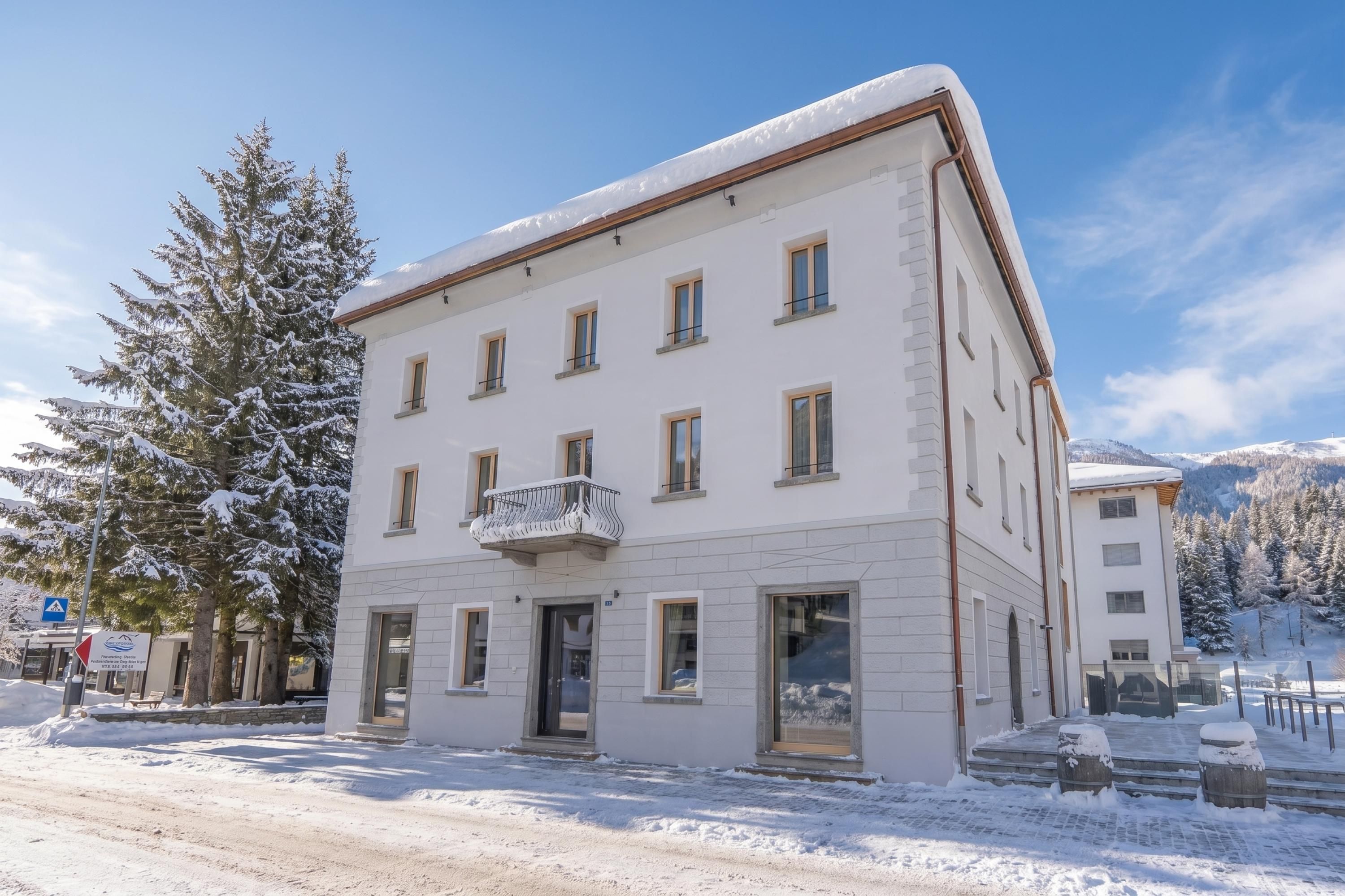 Un edificio de varios pisos con un balcón, rodeado de árboles y montañas cubiertas de nieve.