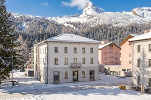 A snow-covered building with a symmetrical facade, large windows, and a flat roof, surrounded by snow-laden trees and mountains. - Residenza Tilia 203 San Bernardino Alpine Apartments, San Bernardino, Switzerland (San Bernardino)