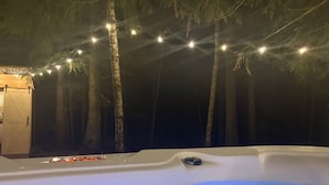 A hot tub with a wooden deck, surrounded by trees and string lights.
