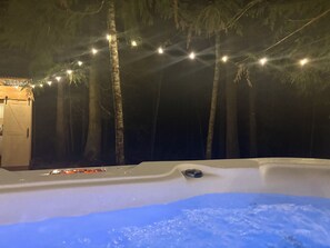 A hot tub with a wooden deck, surrounded by trees and string lights.