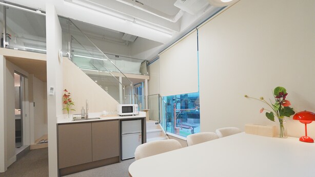 Un espacio de oficina moderno con una mesa de conferencias blanca, una kitchenette, un microondas y vistas al exterior a través de un ventanal grande.