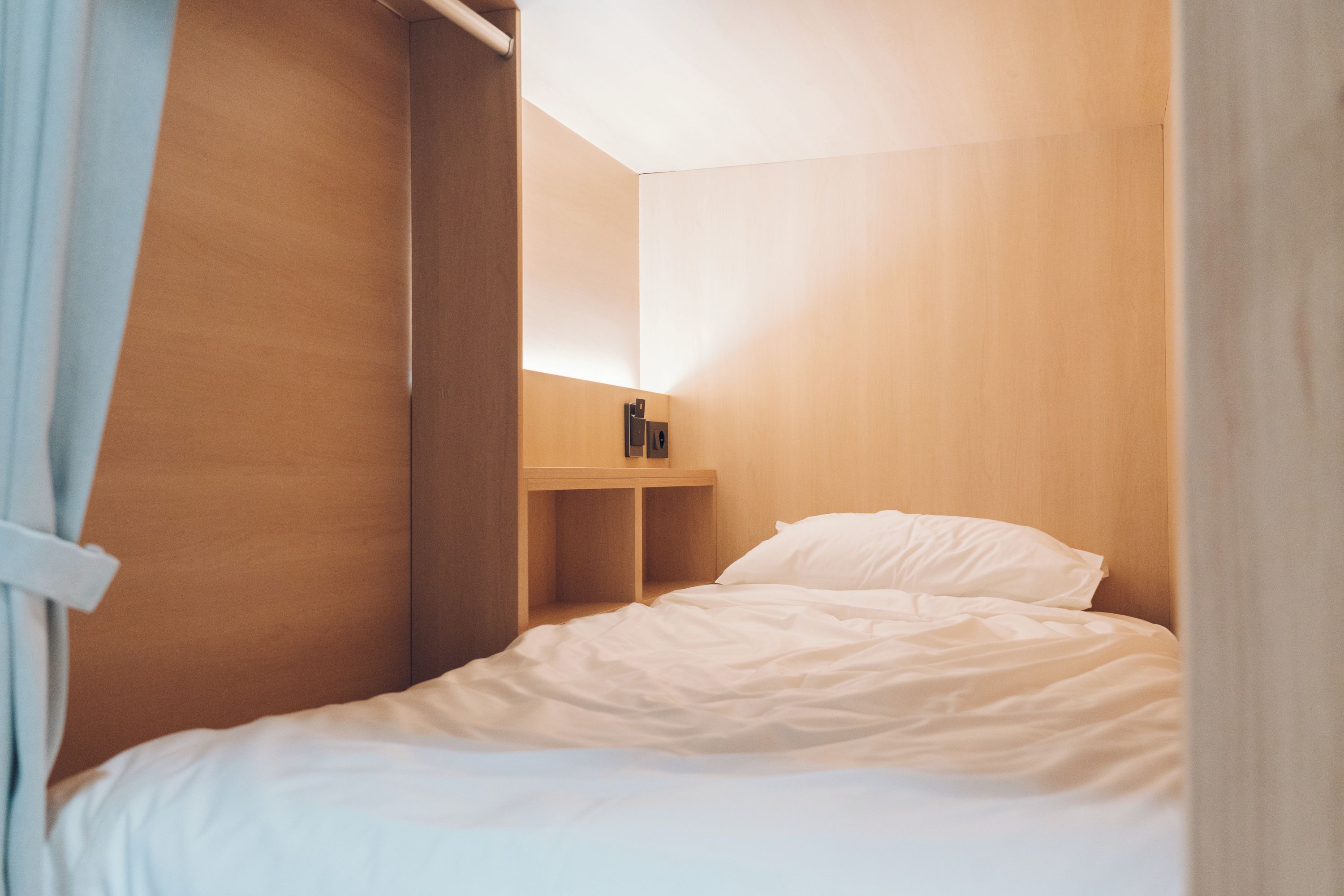 A small, single-bed room with a wooden headboard, a bedside table, and a wall-mounted shelf.