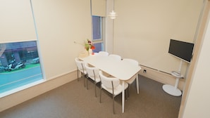 Una habitación con una mesa y sillas blancas, un ventanal pequeño, un televisor montado en la pared y una lámpara en el techo.