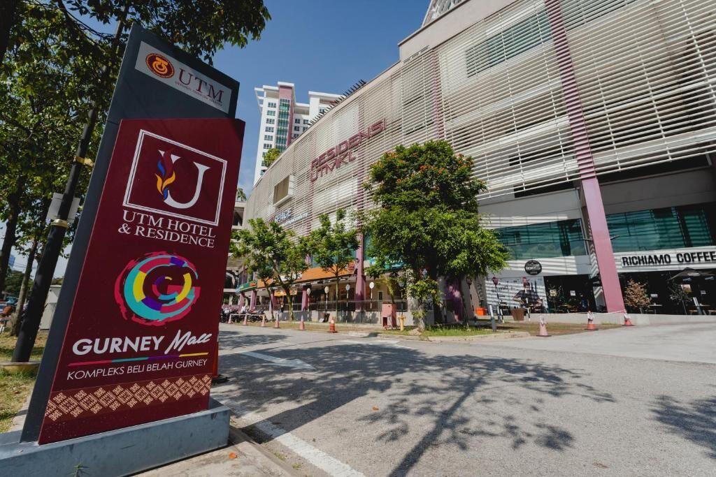 A hotel entrance with a sign for UTM Hotel & Residence, a Gurney Mall, and a modern building with a restaurant.