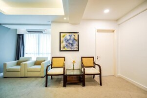 Una habitación con un sofá color beige, dos sillas de madera, una mesa de centro de vidrio y un cuadro abstracto en la pared.