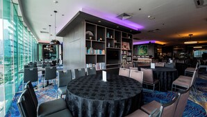A modern library with a large bookshelf, round tables, and chairs, and a view of the city through large windows. - Utm Hotel Residence (Kuala Lumpur)