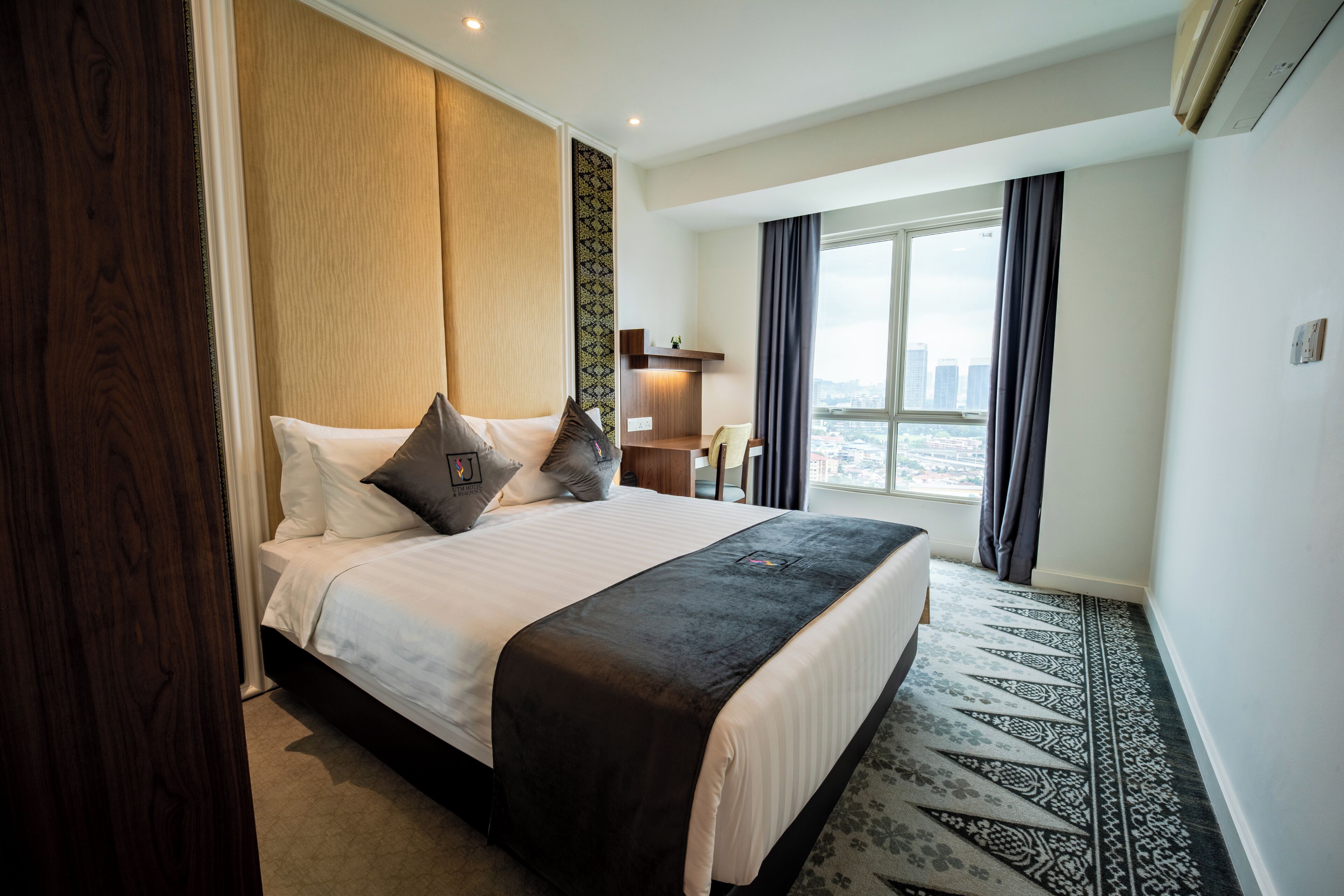 Habitación de hotel con una cama grande, un escritorio, una silla, un ventanal con vista a la ciudad y una alfombra estampada.