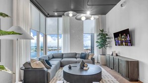 Ein modernes Wohnzimmer mit einem großen, winkelförmigen Schlafsofa, einem runden Couchtisch und Blick auf eine Stadtkulisse durch große Fenster.
