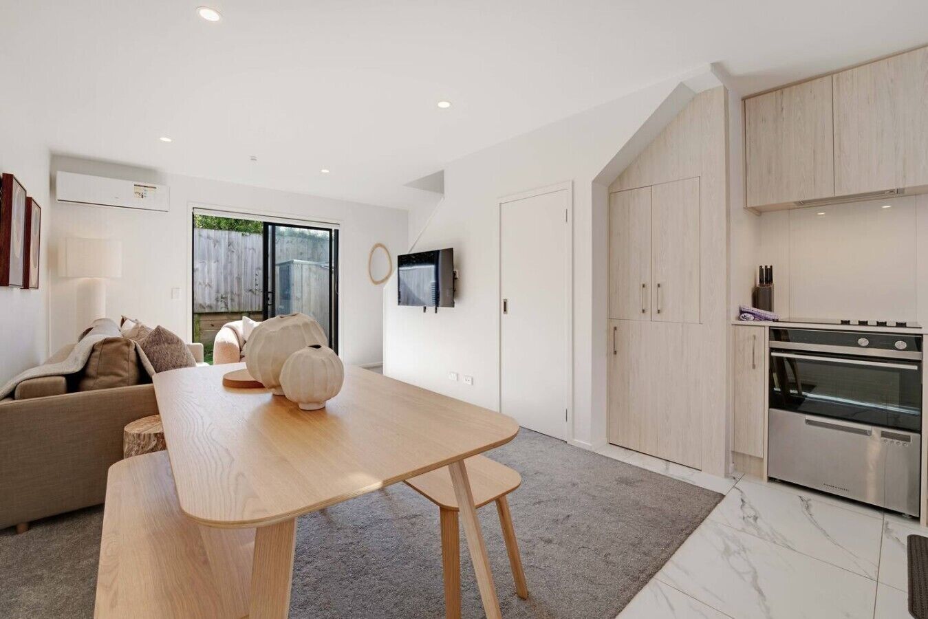 A modern kitchen with a built-in oven and cabinets, a dining area with a wooden table and chairs, and a living room with a sofa and a TV.