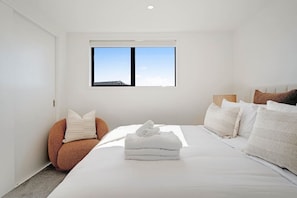 A modern bedroom with a large bed, a single window, and a unique orange chair.