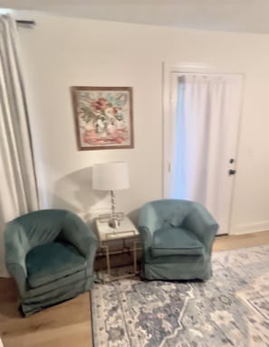 Een kamer met twee fauteuils, een tafeltje met een lamp, een kamerbreed tapijt met dessin en een deur voorzien van gordijnen.