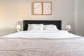 A neatly made bed with a white duvet and pillows, a dark headboard, two bedside lamps, and two framed abstract artworks on the wall. - Blueground | Clarendon, pool, nr Arlington Cemetery (Arlington)