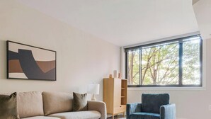 A living room with a sofa, a chair, a bookshelf, and a window with a view of trees. - Blueground | Clarendon, pool, nr Arlington Cemetery (Arlington)