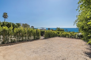Uma área de cascalho com vista para o mar, cercada por vegetação e um portão de metal.