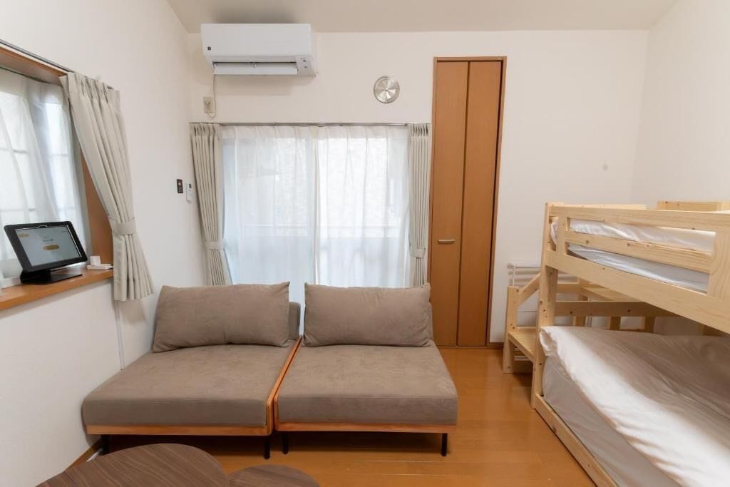 A room with a sofa bed, a wooden bunk bed, a desk with a computer, and a window with curtains.