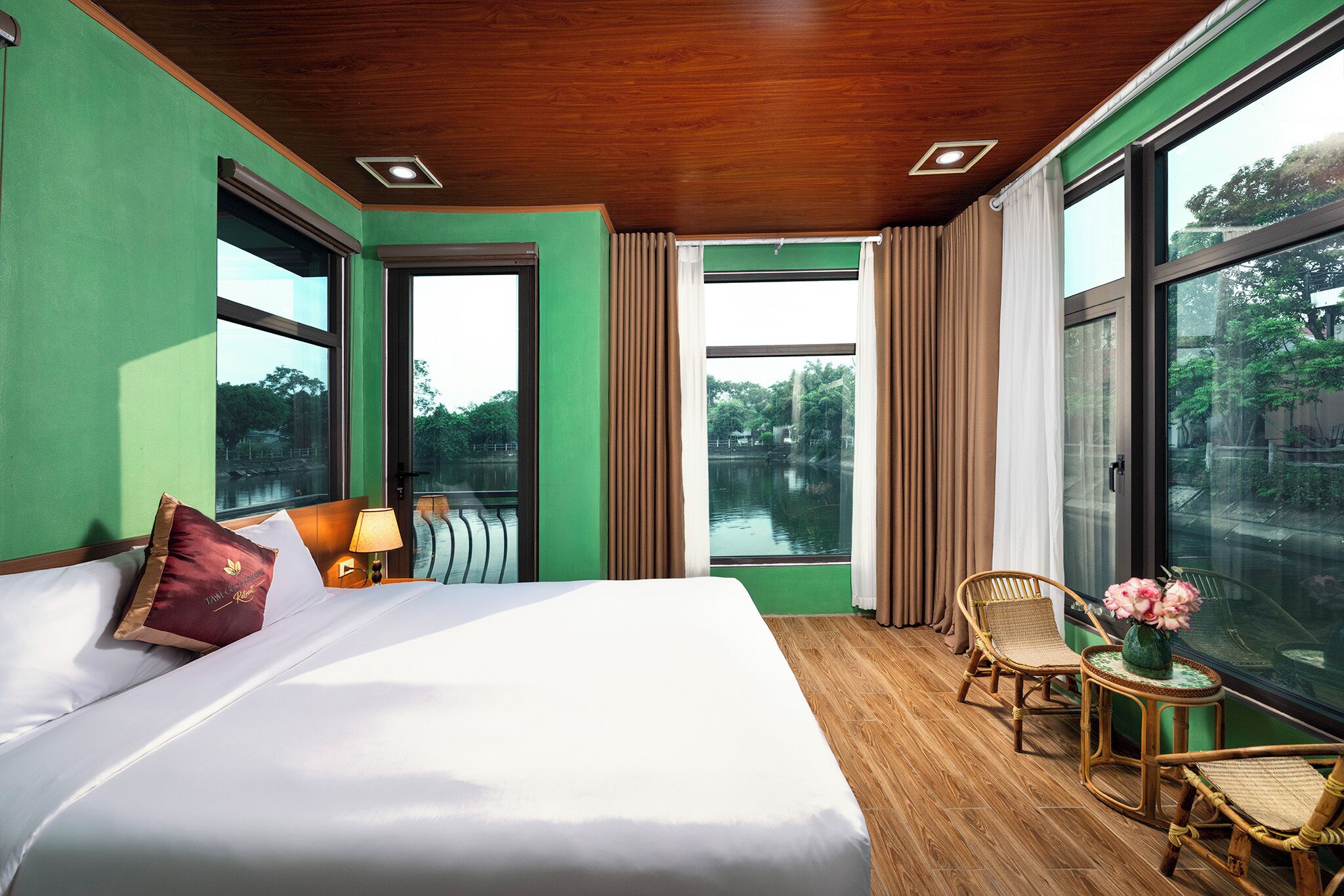 Habitación de hotel con una cama grande, vista a un cuerpo de agua, una mesita con un jarrón de flores y una silla.