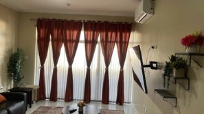 A living room with a sofa, a coffee table, and a TV. There are curtains, a plant, and a wall-mounted shelf with flowers. -  Luxury Escape 1bd/bath apt with pool,balcony  gated complex, 10 mins to airport (Montego Bay)