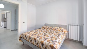 Een slaapkamer met een bed, een radiator en een keuken zichtbaar door een geopende deur.