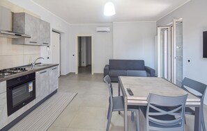 Uma cozinha moderna com fogão a gás, forno e coifa embutida. Há uma área de jantar com mesa e cadeiras, e um sofá cinzento.