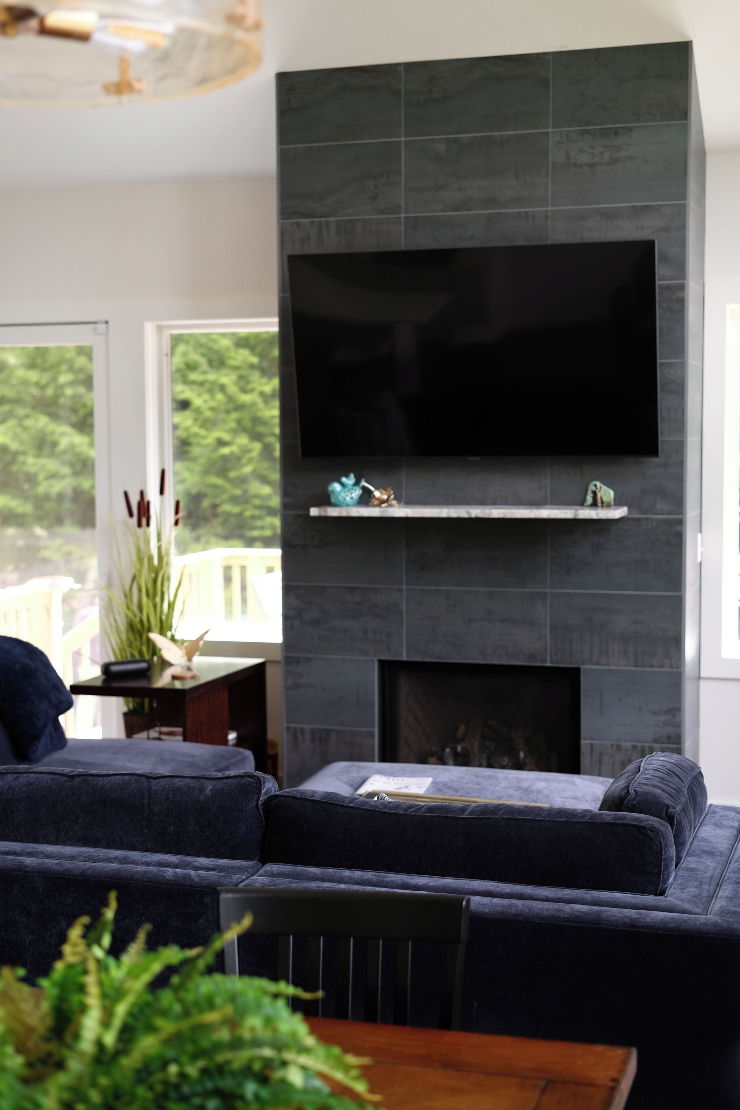 A modern living room with a large flat-screen TV mounted on a dark tiled wall, a sectional sofa, and a small fireplace with decorative items.