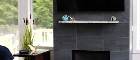A modern living room with a large flat-screen TV mounted on a dark tiled wall, a sectional sofa, and a small fireplace with decorative items.